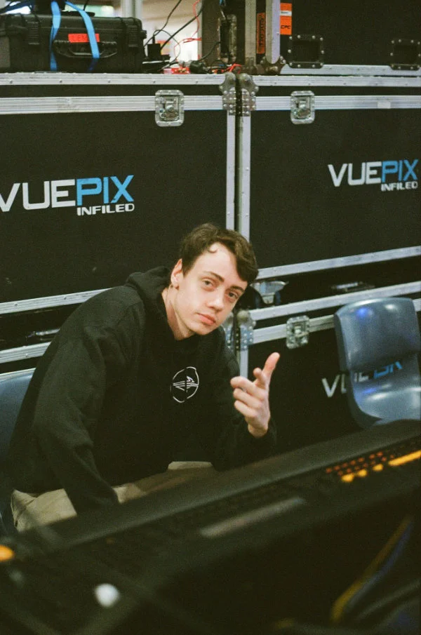 Photograph of a man wearing a black hoodie sitting at a lighting console.